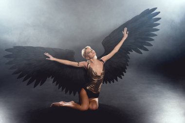 beautiful sexy woman with black angel wings and outstretched hands sitting and posing on dark background clipart