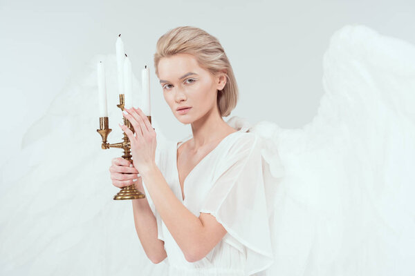 beautiful woman in angel costume with wings holding candelabrum with candles and looking at camera isolated on white
