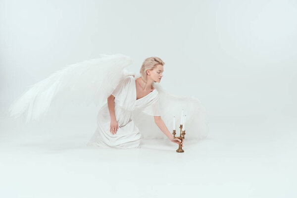 beautiful woman in angel costume with wings sitting near candelabrum with candles on white background