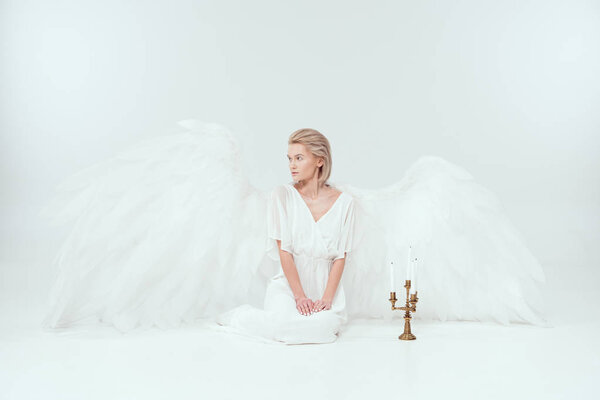 beautiful woman in angel costume with wings sitting near candelabrum with candles and looking away isolated on white