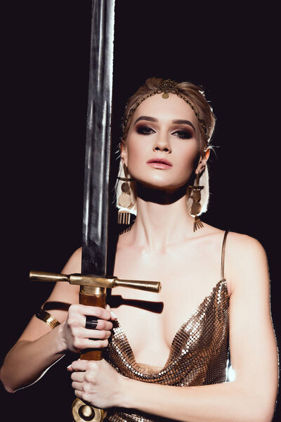 beautiful woman in golden accessories and warrior costume posing with sword isolated on black