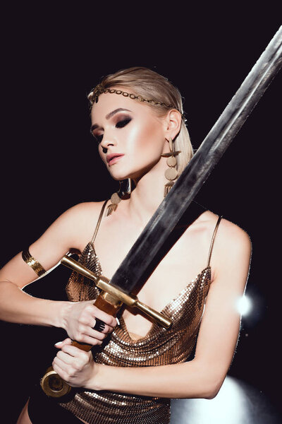 beautiful sexy woman in golden accessories and warrior costume posing with sword on black background