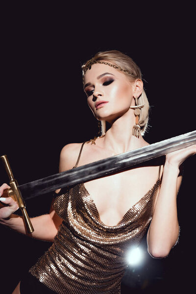 beautiful sexy woman in golden accessories and warrior costume posing with sword on black background