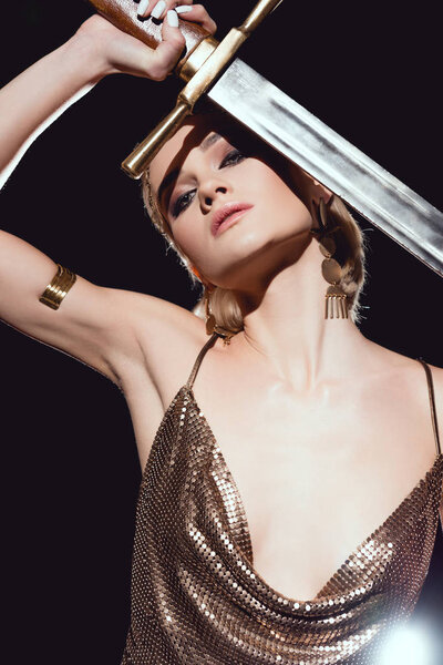 beautiful woman in golden accessories and warrior costume looking at camera and posing with sword on black background