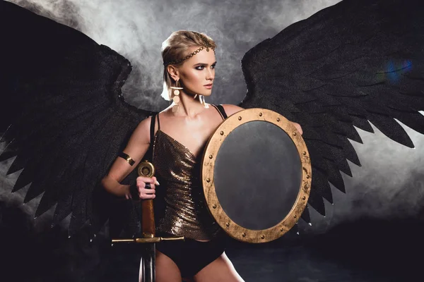 美丽性感的女人在战士服装和天使翅膀摆姿势盾牌和剑在黑色背景 — 图库照片