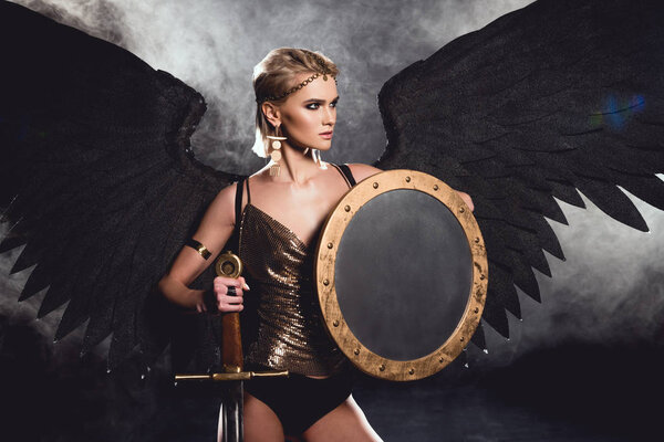 beautiful sexy woman in warrior costume and angel wings posing with shield and sword on black background
