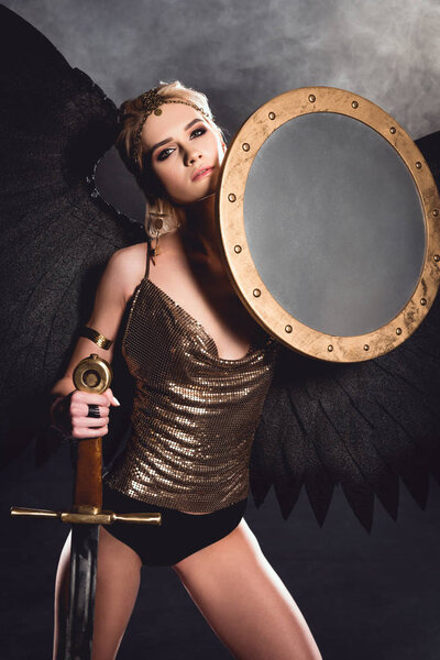 beautiful sexy woman in warrior costume and angel wings posing with shield and sword on dark background