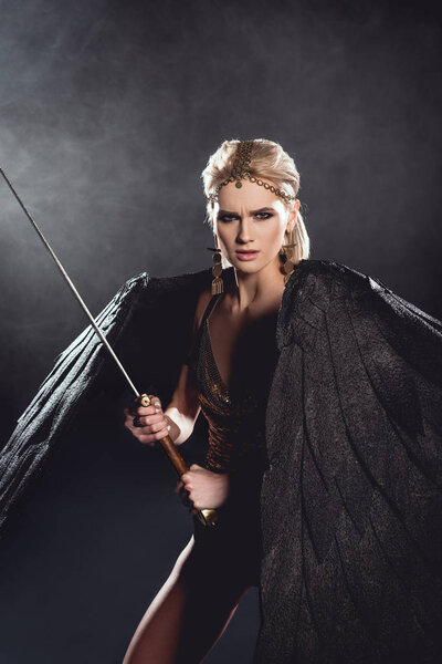 beautiful aggressive woman in warrior costume with angel wings holding sword and looking at camera on black background