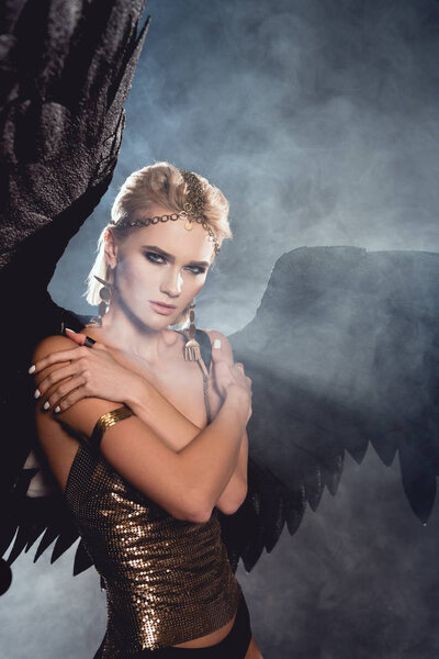 beautiful young sexy woman with black angel wings and golden accessories posing on dark smoky background