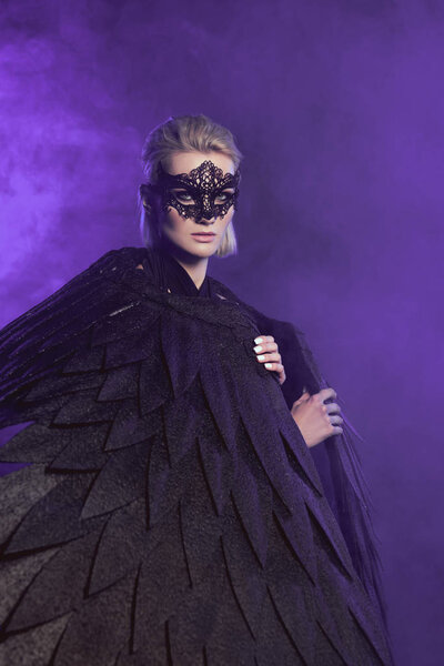 beautiful woman in lace mask and black angel wings looking at camera and posing on violet background