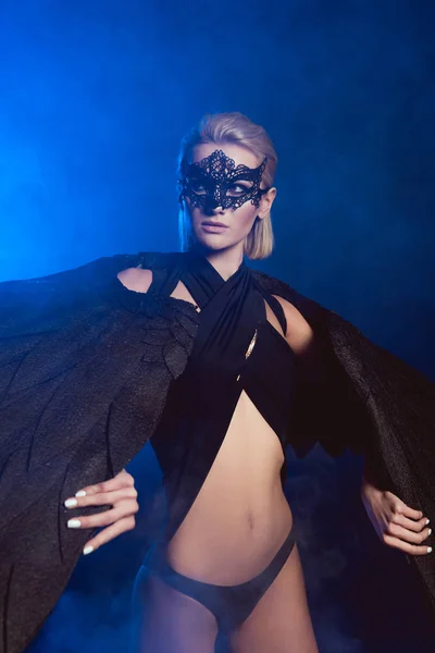 美丽性感的女人在蕾丝面具和黑色天使翅膀摆姿势和看着深蓝色的背景 — 图库照片