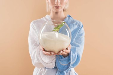 cropped view of woman holding fish bowl with sand and small green plant  while standing in eco clothing isolated on beige, environmental saving concept  clipart