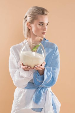 beautiful young woman holding fish bowl with sand and small green plant  while standing in eco clothing isolated on beige, environmental saving concept  clipart