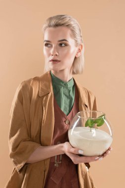 blonde woman holding fish bowl with sand and small green plant while standing in eco clothing isolated on beige, environmental saving concept  clipart