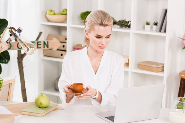 attractive woman looking at laptop while holding cup in office 