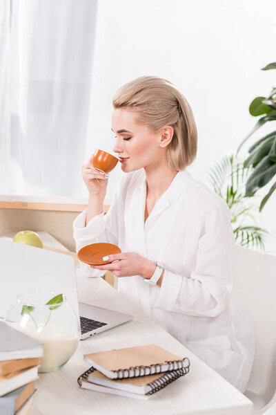 attractive woman drinking coffee from cup near laptop in office 