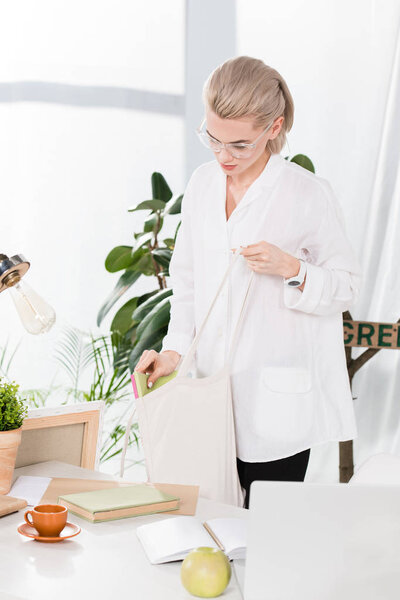 beautiful woman in glasses holding eco bag near desk in office, environmental saving concept 