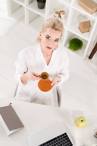 overhead view of woman in glasses holding cup of coffee near desk in office