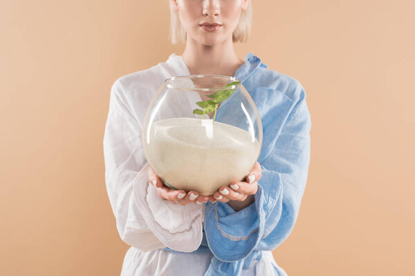 cropped view of woman holding fish bowl with sand and small green plant  while standing in eco clothing isolated on beige, environmental saving concept 