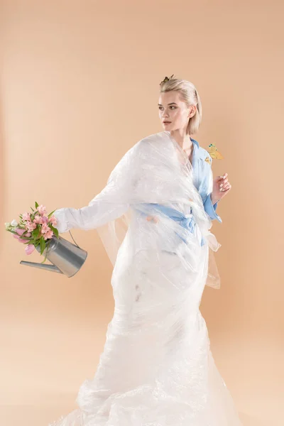 ベージュ 環境保存の概念に分離された花の水まき缶を押しながらエコ服で立っているポリエチレンに包まれた美しい少女 — ストック写真