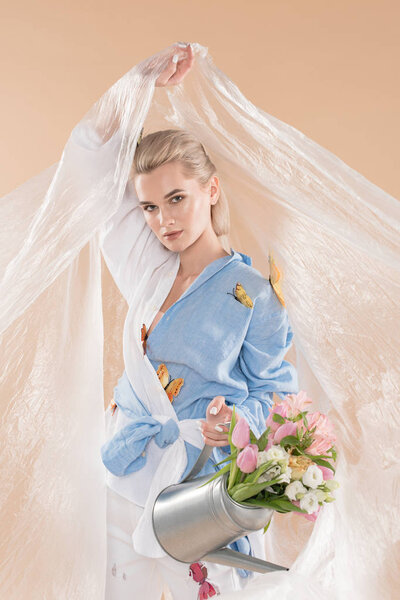 young woman holding polyethylene and watering can with flowers while standing in eco clothing isolated on beige, environmental saving concept 