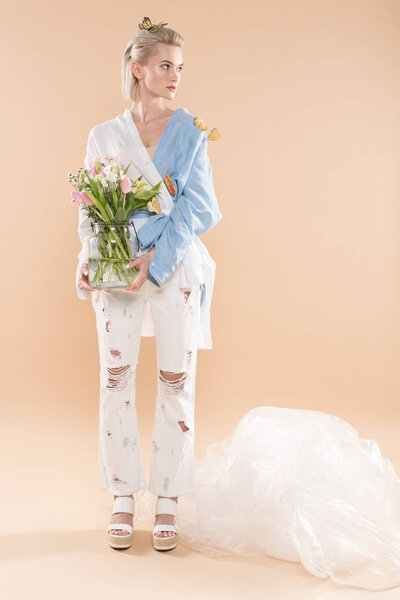 beautiful yound woman standing with butterflies on eco clothing and holding glass vase with flowers on beige background, environmental saving concept