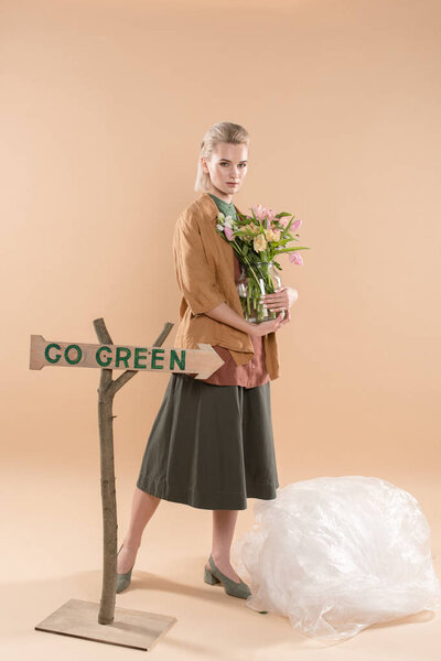 attractive girl in eco clothing holding vase with flowers near sign with go green lettering on beige background, environmental saving concept