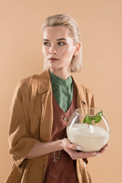 blonde woman holding fish bowl with sand and small green plant while standing in eco clothing isolated on beige, environmental saving concept 