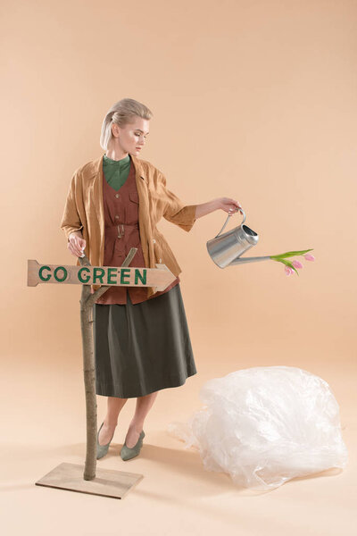привлекательная девушка в экологической одежде проведение полива банка с цветами рядом с табличкой с идти зеленые буквы на бежевом фоне, концепция охраны окружающей среды
