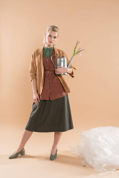 attractive girl in eco clothing holding watering can with flowers near polyethylene on beige background, environmental saving concept