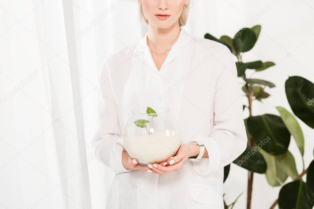 cropped view of woman holding glass fish bowl with sand near and green leaves,  environmental saving concept 