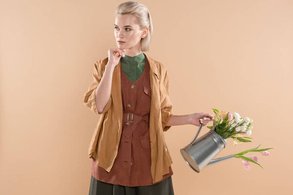 pensive girl in eco clothing holding watering can with flowers isolated on beige background, environmental saving concept