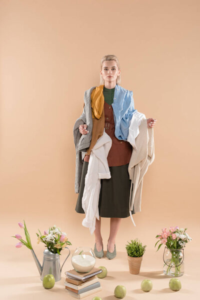 blonde woman holding eco clothing near plants and flowers on beige background, environmental saving concept