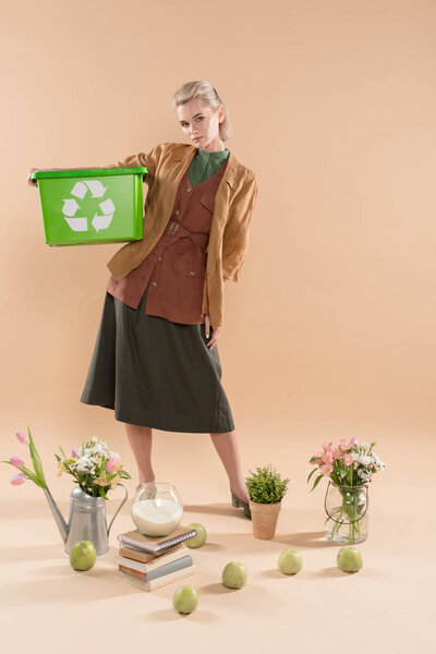 beautiful woman holding recycling box near plants and flowers on beige background, environmental saving concept