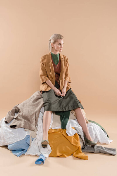 blonde woman sitting on stack of clothing on beige background, environmental saving concept