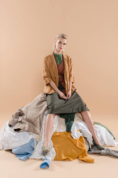 blonde woman sitting on pile of clothing on beige background, environmental saving concept