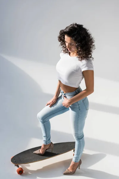 Curly woman in jeans and crop top standing on longboard on grey background