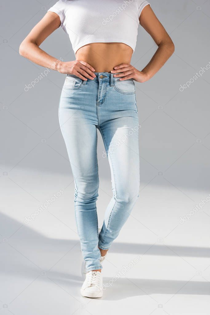 Cropped view of girl in jeans posing with arms akimbo on grey background