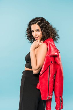 Graceful curly girl holding red leather jacket on blue background