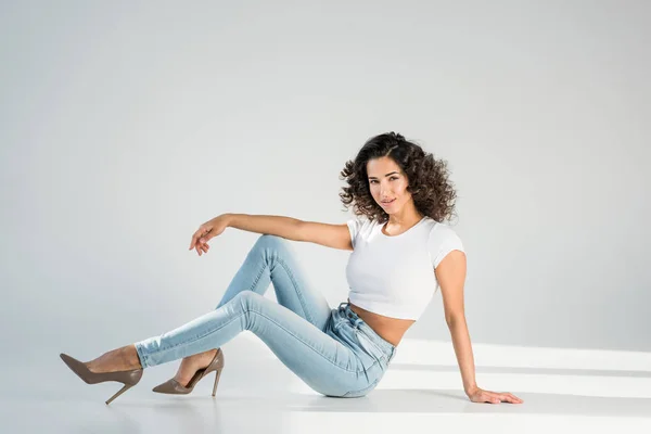 Stylish woman in jeans and high-heeled shoes sitting on grey background