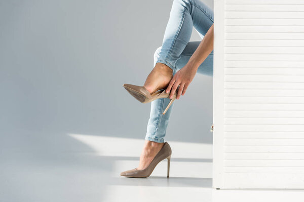 Partial view of woman putting on high-heeled shoes behind room divider on grey background