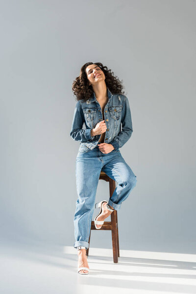Blissful young woman in jeans and jacket sitting on wooden chair on grey background