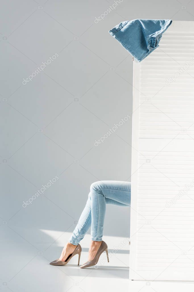 Partial view of woman in jeans sitting behind room divider on grey background