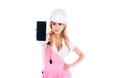 blonde handy woman in pink overalls, hardhat standing and holding smartphone with blank screen isolated on white clipart