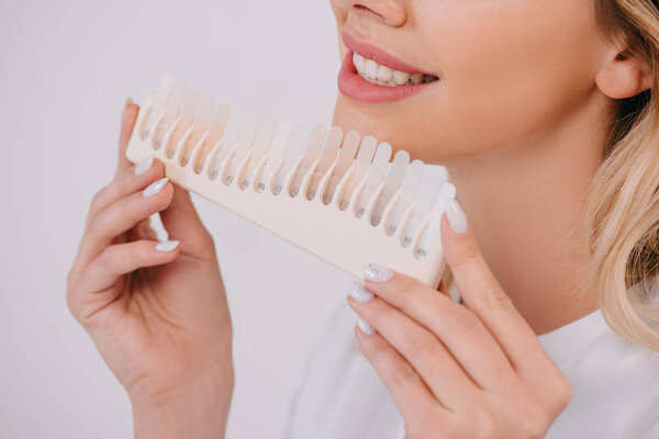 partial view of smiling woman holding teeth color palette isolated on white, teeth whitening concept