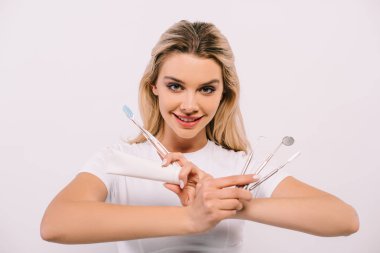 beautiful smiling woman looking at camera while holding toothpaste, toothbrush and dental instruments isolated on white clipart