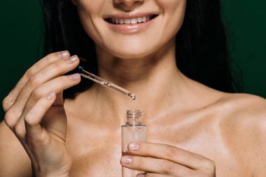 cropped view of smiling naked woman holding pipette and bottle with serum isolated on green clipart