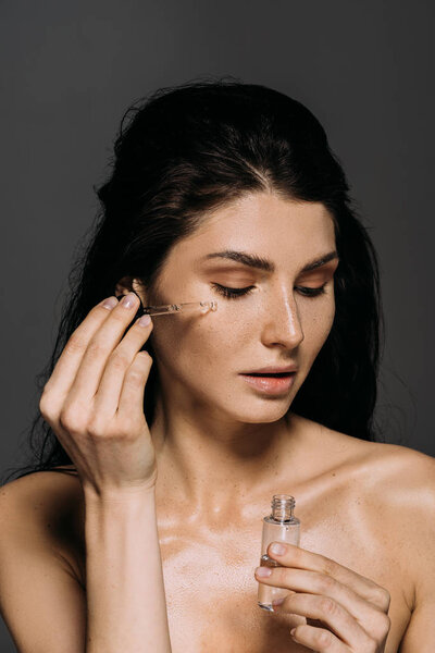 naked brunette girl applying serum on face with pipette isolated on grey