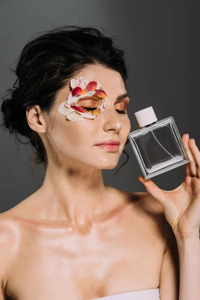 tender girl with closed eyes and petals on face holding perfume bottle isolated on grey