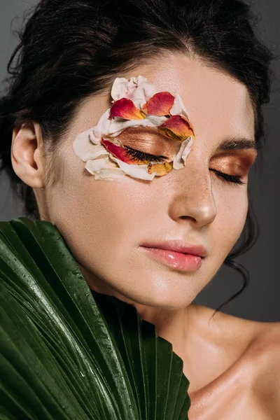 温柔的女孩与闭上的眼睛与化妆和花卉花瓣在脸上举行着热带叶子隔离在灰色 — 图库照片